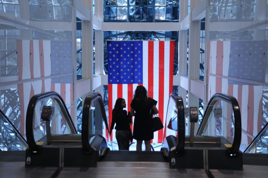 Dve žene se spuštaju pokretnim stepenicama u zgradi u kojoj je zakačena zastava SAD-a