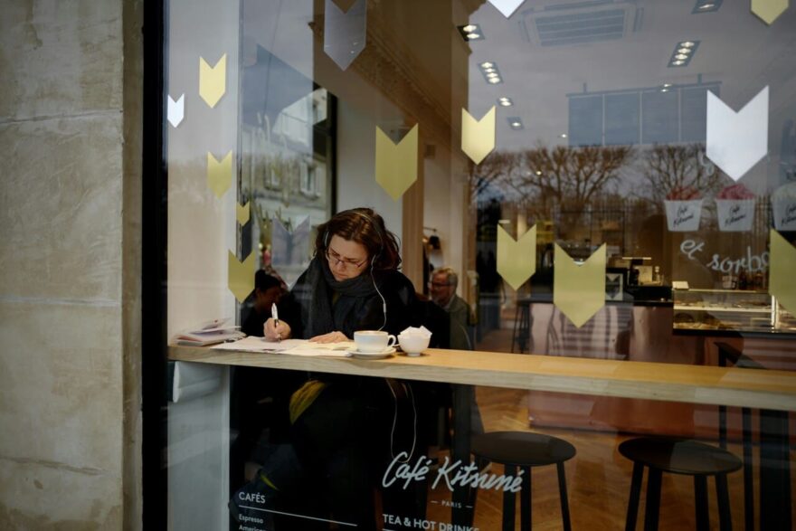 Žena sa slušalicama sedi pored izloga kafića i piše u svesci uz šolju kafe
